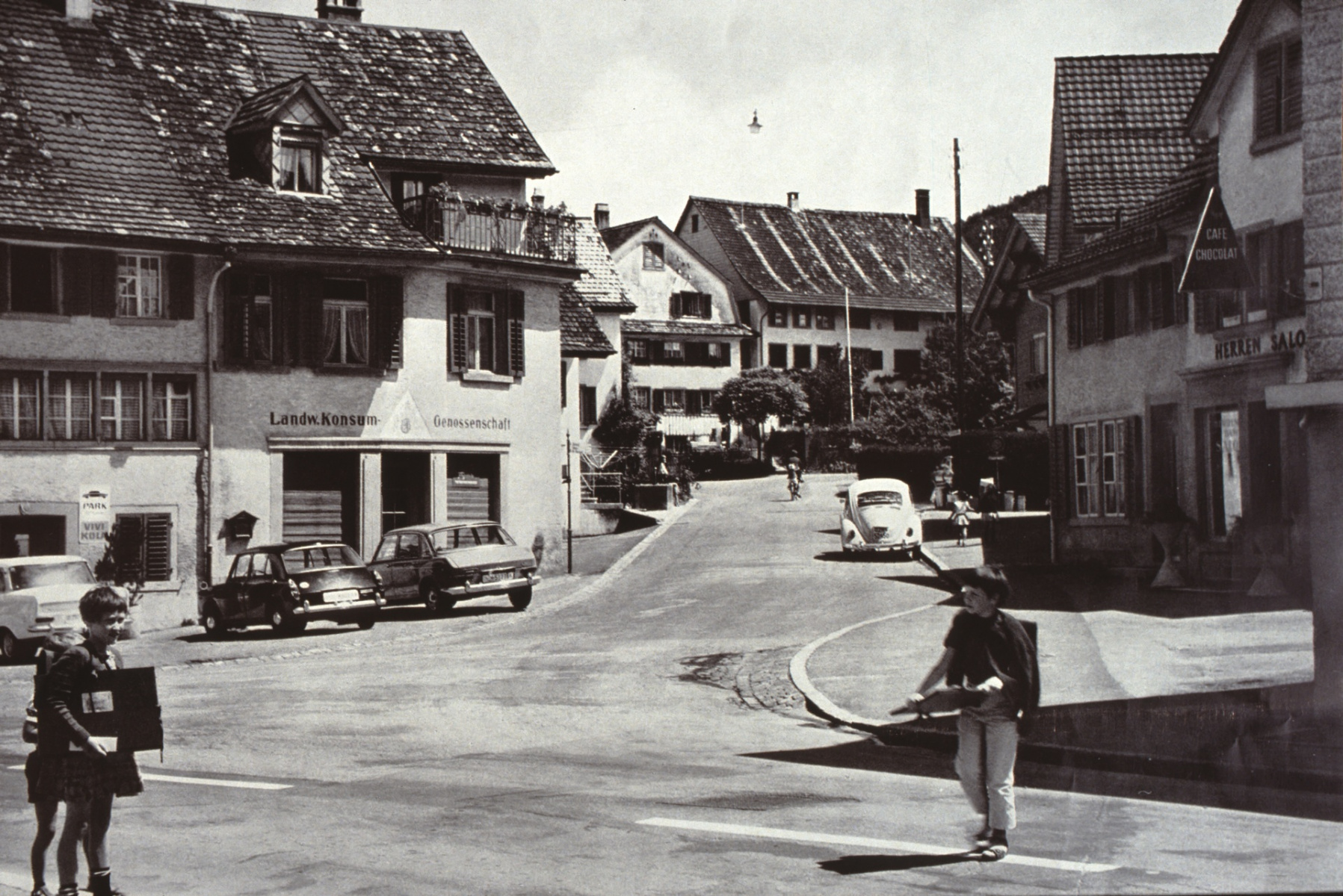 Bärenplatz, Bettswilerstr. Auch in Bäretswil hält das Auto um 1960 breiteren Einzug. Auf der Strasse nach Bettswil ist eine Mutter mit ihrem Töffli und dem Kind auf dem Gepäckträger zu erkennen.
