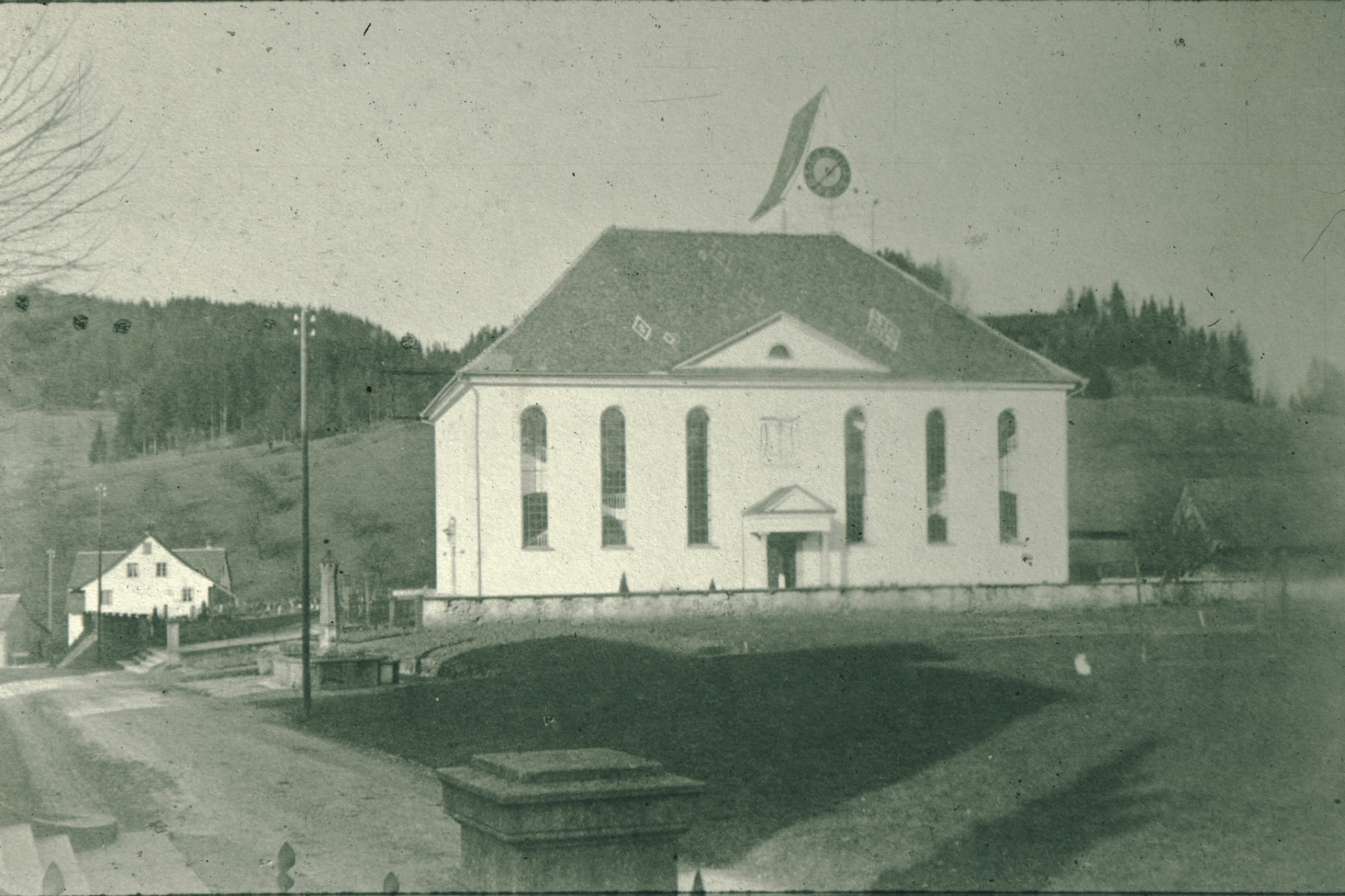 Ref. Kirche, kleiner Friedhof, Eingänge. Bis 1927 hat nur der Haupteingang Süd ein Vordach. Die Telefonstangen erinnern an erste Telefone, die es in Bäretswil seit 1887 gibt.